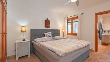 Un dormitorio con cama, mesitas de noche, ventilador de techo, ventana con cortinas y un baño visible a través de una puerta abierta.