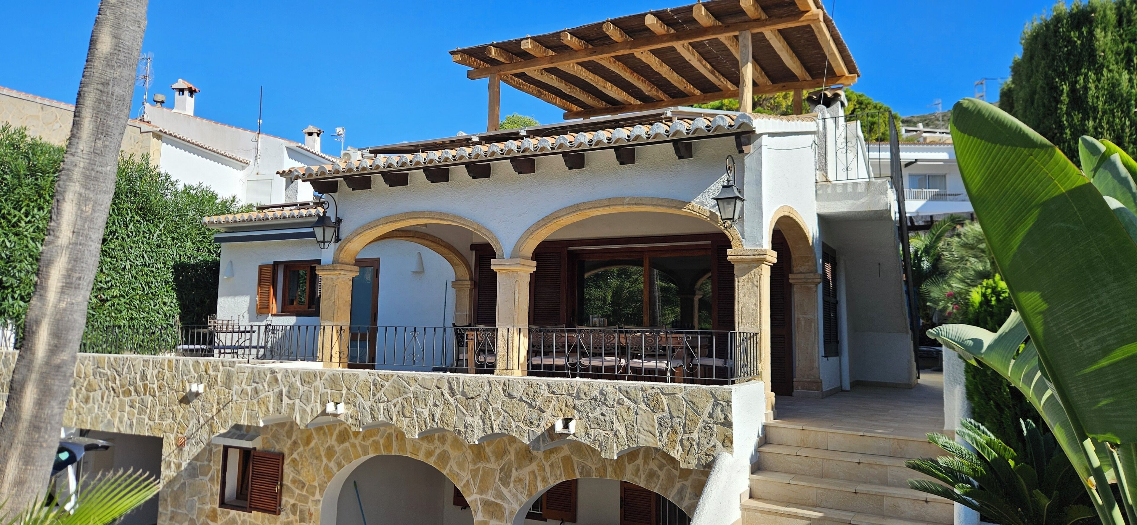 A two-story building with a stone facade, a balcony with a wooden pergola, and a view of the surrounding greenery.