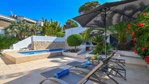 A pool area with lounge chairs and a large umbrella, surrounded by greenery and a stone wall.