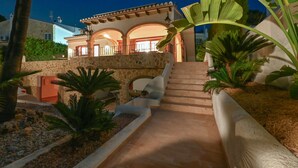 A stone staircase leading to a house with a balcony, surrounded by palm trees and a garden.