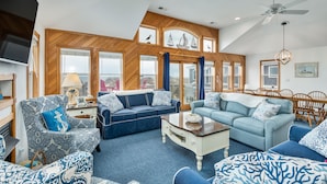 Ein geräumiges Wohnzimmer mit hohen Decken, Holzvertäfelung und großen Fenstern. Es gibt zwei Sofas, einen Couchtisch und einen Fernseher.