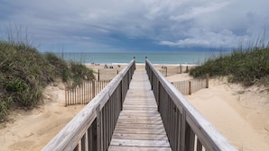 Ein hölzerner Steg führt zu einem Strand mit Sand und Dünen an beiden Seiten.