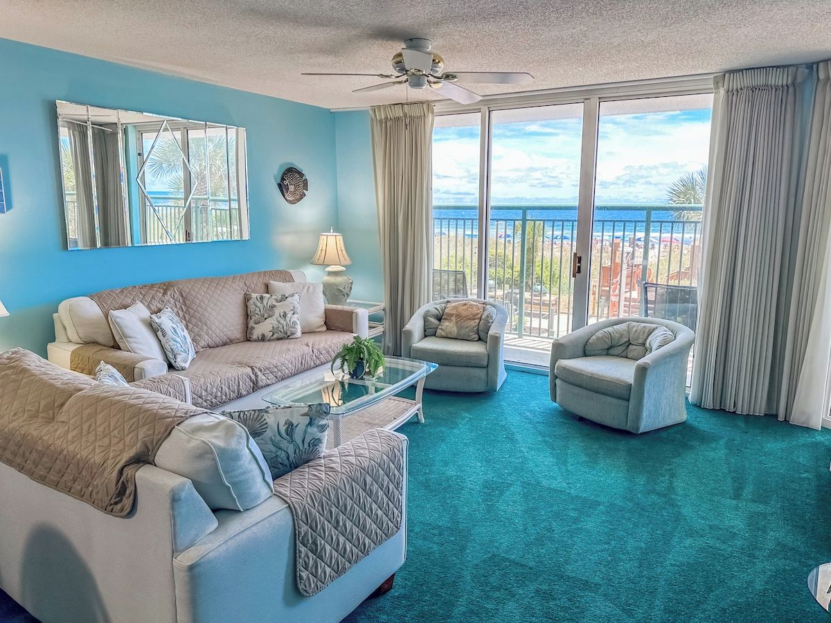 A living room with a large window, a ceiling fan, and a view of the ocean.