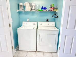 A laundry room with two white front-loading washers and dryers, a shelf with various items, and a door on the left.