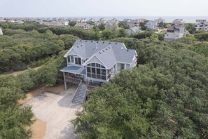 A large house with a covered porch and a staircase leading to a deck, surrounded by dense greenery.