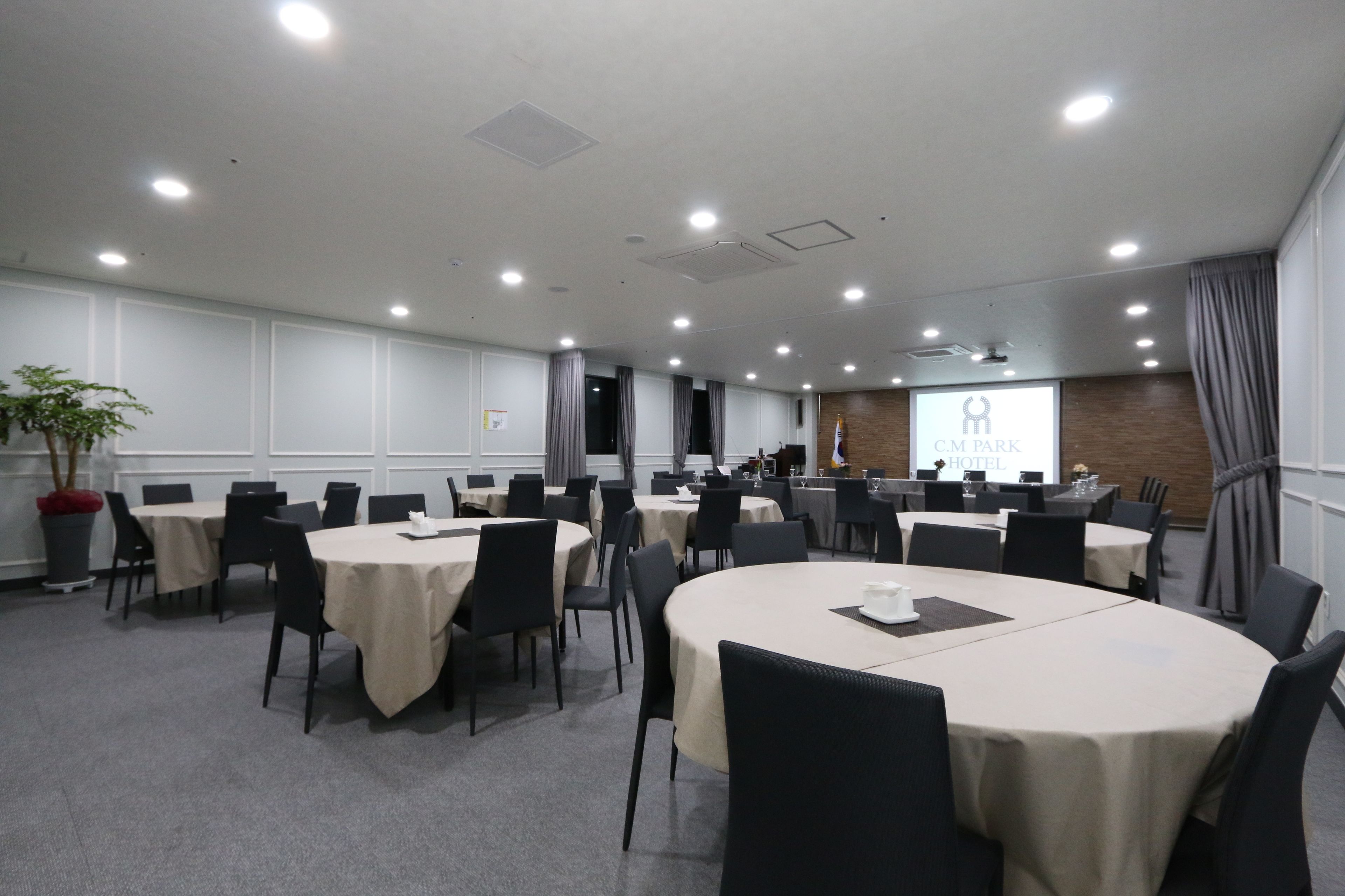 Un salón de conferencias con mesas redondas, sillas y una pantalla de proyección que muestra el logotipo de un hotel.