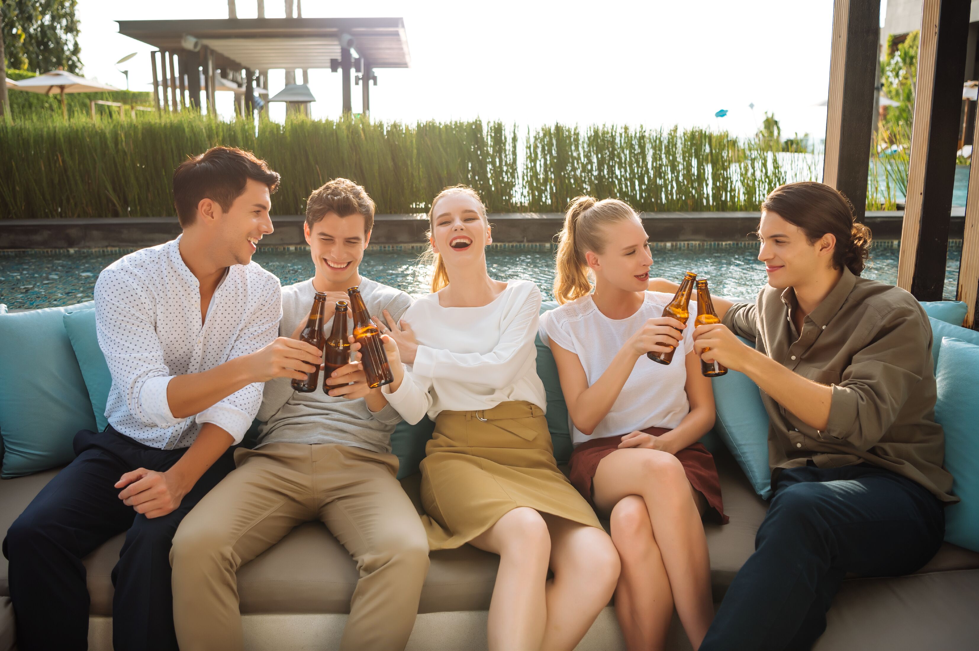 Een groep mensen zit op een bank bij een zwembad, met drankjes in hun handen.