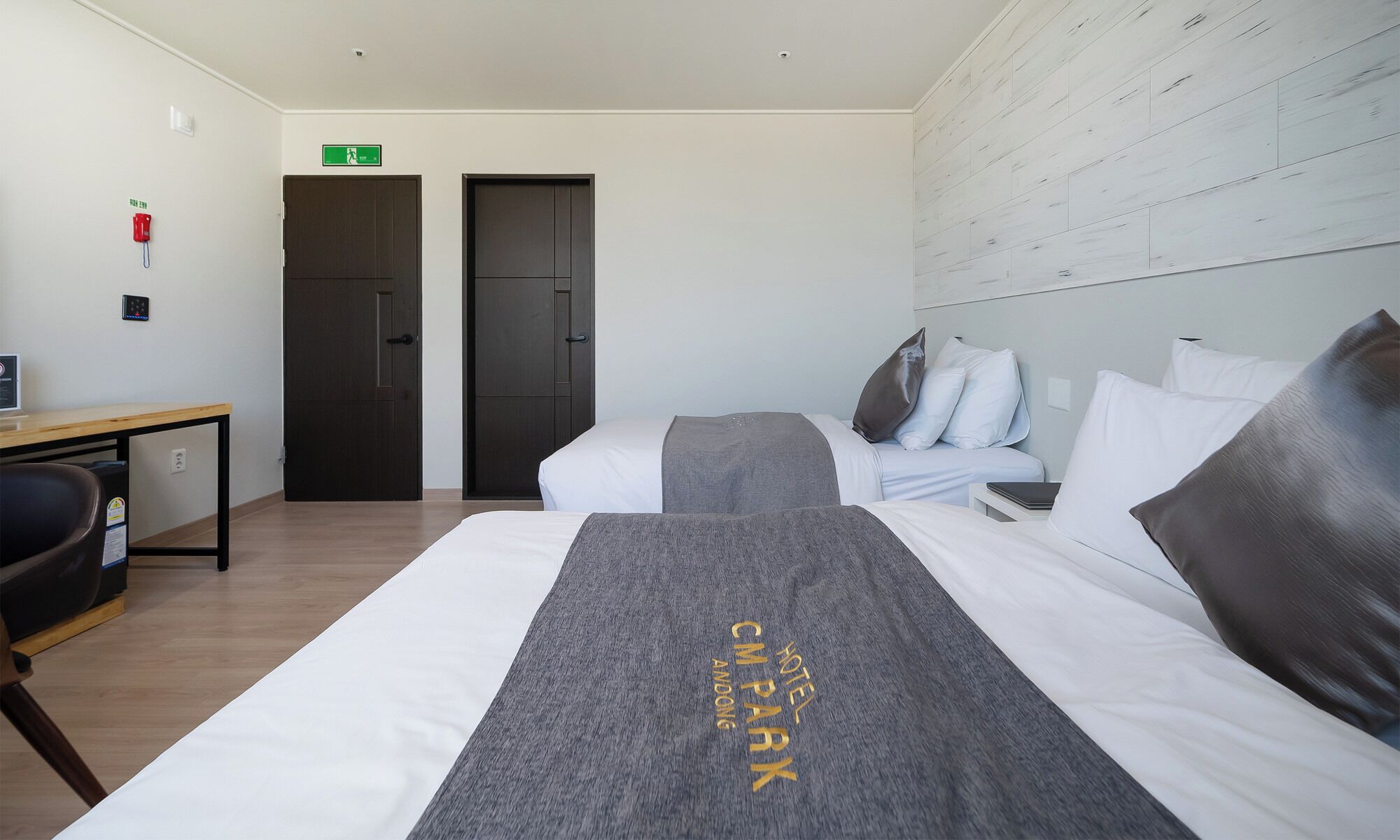 Une chambre d’hôtel avec deux lits, un bureau et une chaise. On y trouve une porte avec un panneau indiquant la sortie de secours, un téléphone et un interrupteur sur le mur.