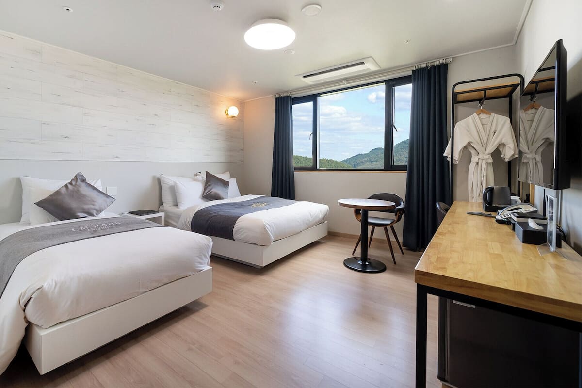 Habitación de hotel con dos camas, un escritorio, una silla, un televisor y una ventana con vistas a las colinas.