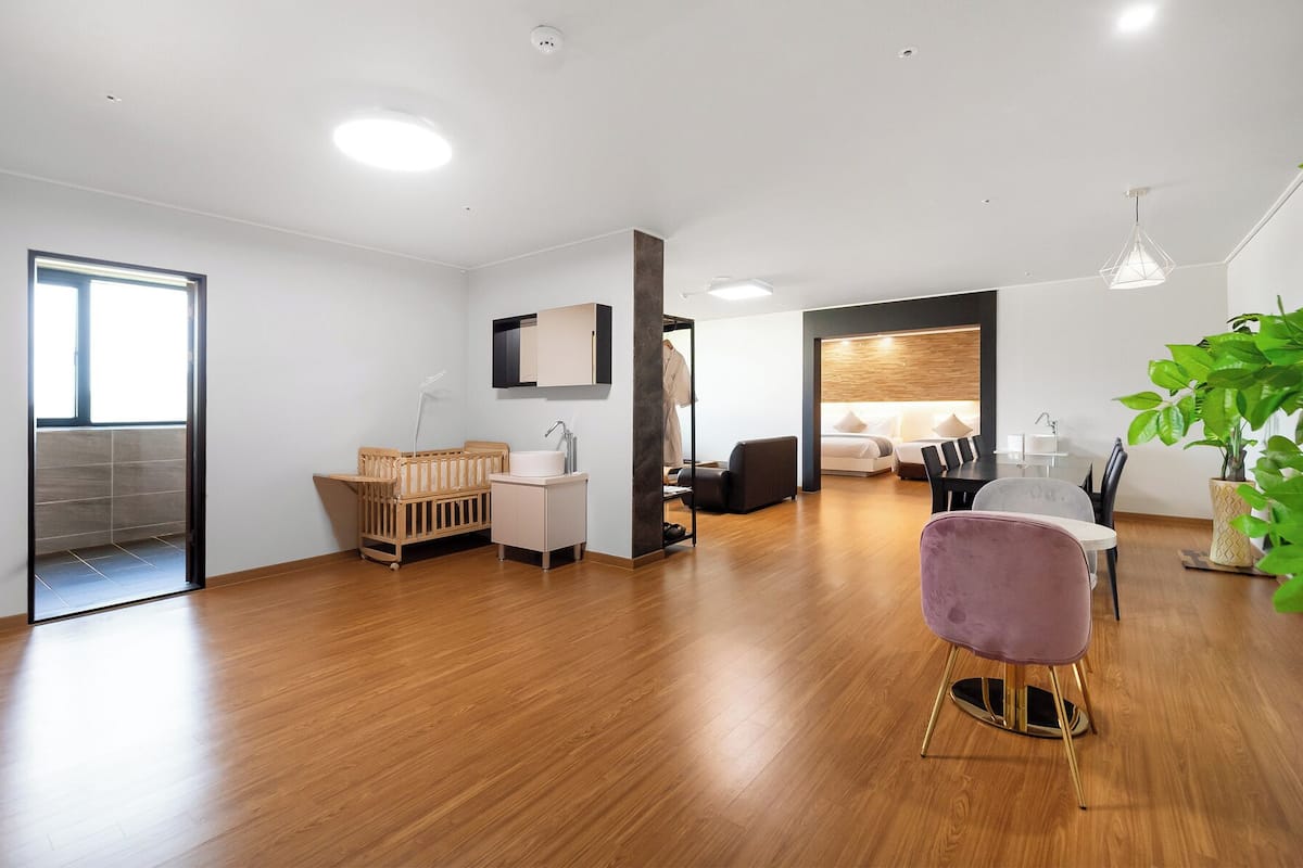 Una habitación de hotel moderna con suelo de madera, una zona de comedor con mesa y sillas, un ventanal grande y una planta.