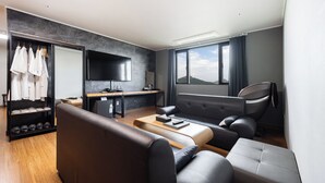 A modern hotel room with a sofa, a small dining table, a flat-screen TV, a wardrobe with hanging clothes, and a window with a view of the mountains. - Andong CM Park Hotel (Yecheon)