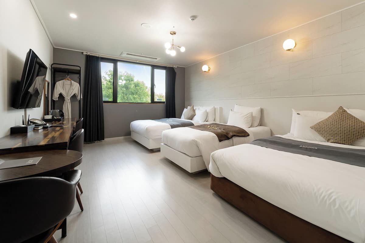 Habitación de hotel con dos camas, un escritorio, un televisor y una ventana con vistas a árboles.