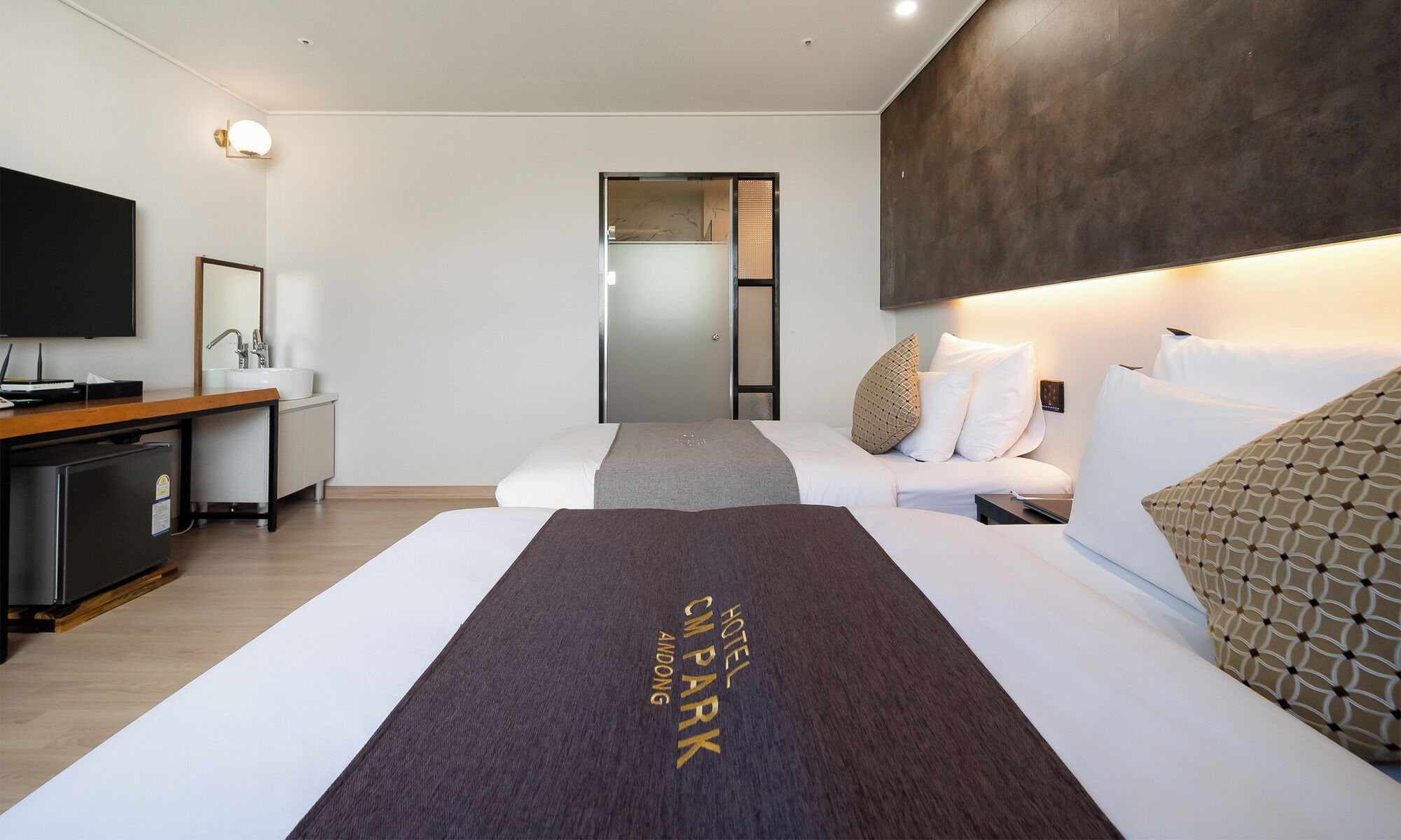 A hotel room with a bed, a desk, a television, and a bathroom visible through a glass door.