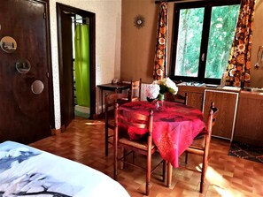 A room with a wooden floor, a dining table with a red tablecloth, a chair, a vase with flowers, a window with curtains, and a door with a mirror and a light switch.