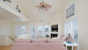 Ein großzügiger Wohnraum mit einem großen rosa Sofa, einem Deckenventilator, einem Kamin und einem Fernseher auf einem weißen Standfuß.