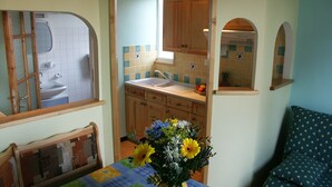 Une cuisine de taille réduite, avec des meubles en bois, un évier et une fenêtre. Une table agrémentée d’un arrangement floral se trouve au premier plan.