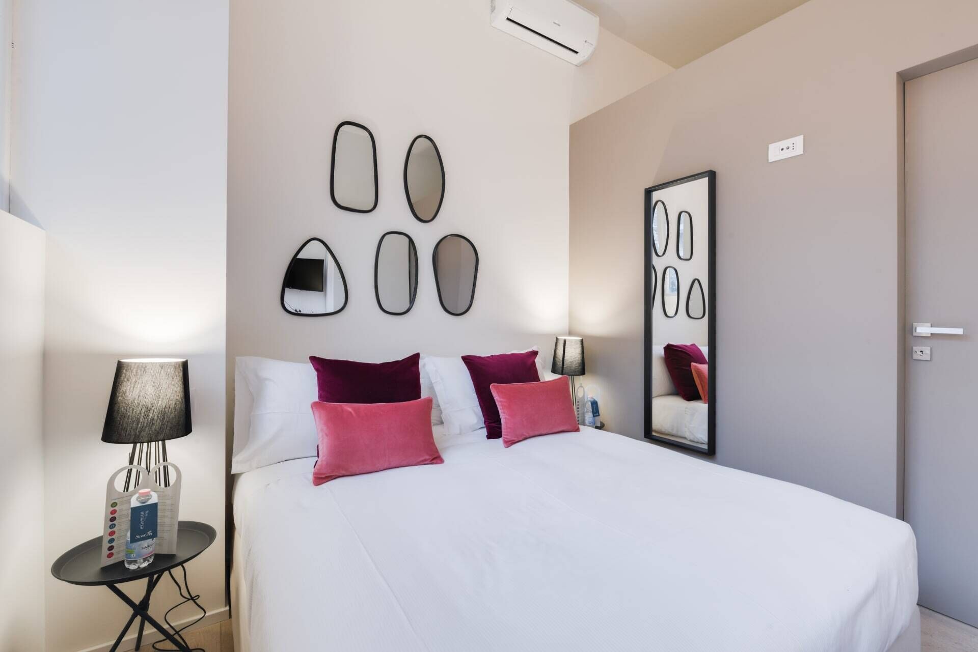 Ein Hotelzimmer mit Bett, Nachttisch mit Lampe, Wasserflasche und einer Wand mit abstrakten Spiegeln.