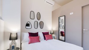 Una habitación de hotel con una cama, una mesita de noche con lámpara, una botella de agua y un espejo de pared abstracto.