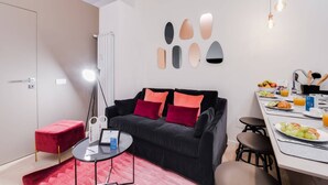 Una sala de estar moderna con un sofá negro, cojines rojos, una mesa de centro de vidrio y un reposapiés rojo. Hay una zona de comedor con una mesa blanca, sillas y un frutero.