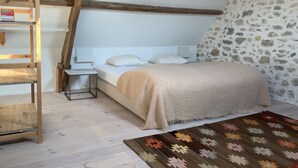Een slaapkamer met een schuine kap, een bed met een beige deken, een houten trap, een klein tafeltje en een kamerbreed tapijt met een patroon.