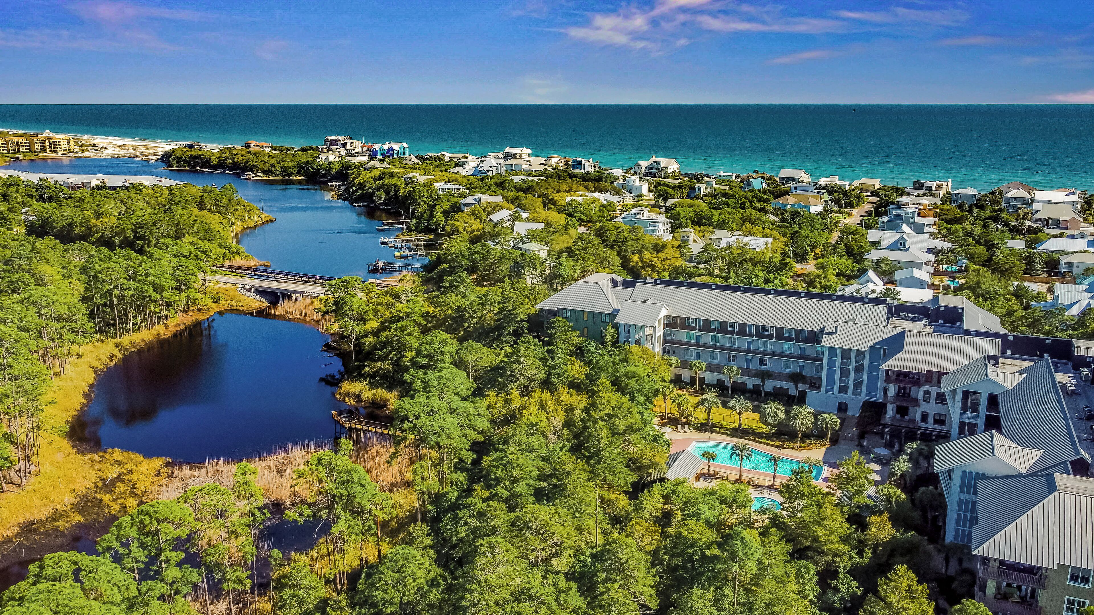 An aerial view of a coastal resort with a large building featuring a swimming pool, surrounded by lush greenery and a clear view of the ocean.