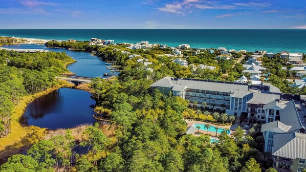 An aerial view of a coastal resort with a large building featuring a swimming pool, surrounded by lush greenery and a clear view of the ocean.