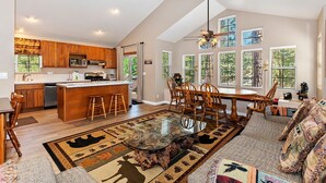 A spacious living area with a large glass coffee table, wooden furniture, and a view of the outdoors through large windows.