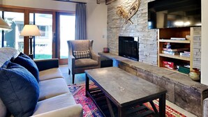 A living room with a stone fireplace, a sofa, a coffee table, and a television mounted on the wall. - 🌲Timberline C1A Deluxe 3-Bedroom ~Hot Tub~Pool~WiFi~Ski-in/out~Local Shuttle🌲 (Snowmass Village)