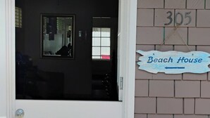 Eine weiße Tür mit Fußmatte, ein Schild mit der Aufschrift 'Beach House' darüber und eine Ziegelwand mit der Nummer '305'.