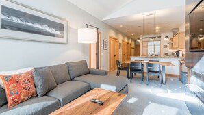 Un salon moderne avec un canapé gris, une table basse en bois et un coussin décoratif aux couleurs vives. On y trouve également un coin repas avec une table et des chaises en bois, ainsi qu’une cuisine dotée de meubles en bois et d’un réfrigérateur en acier inoxydable.