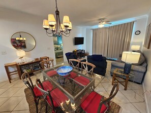 A living room with a glass-top coffee table, red cushioned chairs, a dark sofa, a television, and a ceiling fan.