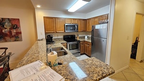 A kitchen with granite countertops, wooden cabinets, a stainless steel refrigerator, a microwave, and an oven. - The Inn at Dauphin Island 104 Walk right out to Pools and Beach! (Dauphin Island)