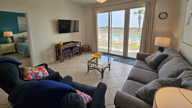 A living room with a sofa, a television, a dining table, and a view of a pool. - The Inn at Dauphin Island 104 Walk right out to Pools and Beach! (Dauphin Island)