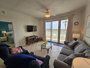 A living room with a sofa, a television, a dining table, and a view of a pool.