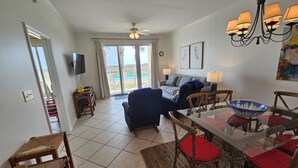 A living room with a TV, a sofa, a dining table with chairs, and a balcony with a pool view. - The Inn at Dauphin Island 104 Walk right out to Pools and Beach! (Dauphin Island)