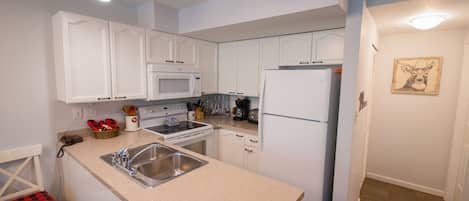 Una cocina compacta con gabinetes blancos, microondas, estufa, fregadero y refrigerador.