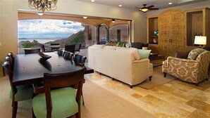 A spacious living area with a large dining table, a sofa, and a patterned armchair. There is a view of the outdoors through an open archway.