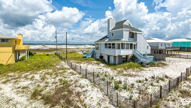 A beachfront house with a staircase leading to the main entrance, surrounded by a wooden fence and sandy terrain. - “Sea La Vie” Water Views,  Steps to the beach (Navarre Beach)