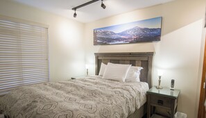 Un dormitorio con cama, mesita de noche, lámpara y un cuadro de un paisaje montañoso.