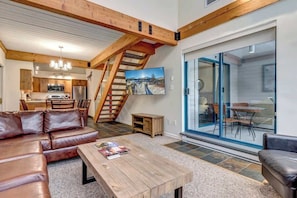 A spacious living area with a wooden ceiling, a sofa, a coffee table, a kitchen in the background, and a staircase leading to an upper level.