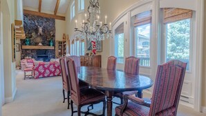 A dining area with a large round table, six chairs, and a chandelier. There is a stone fireplace and a kitchen in the background. - Lodge Apt. II 886 - Perfect for Families Sun Valley Resort Style & Amenities (Sun Valley)