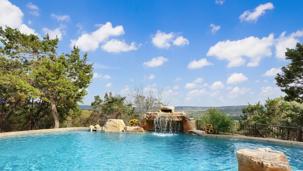 Une piscine d’un bleu éclatant, agrémentée d’une cascade, entourée d’arbres et offrant une vue sur des collines lointaines.