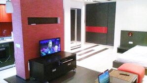 Ein modernes Hotelzimmer mit Flachbildfernseher, einem Laptop auf einem Holztisch, einem Bett mit roter Bettwäsche und einem separaten Schlafbereich mit Kleiderschrank.