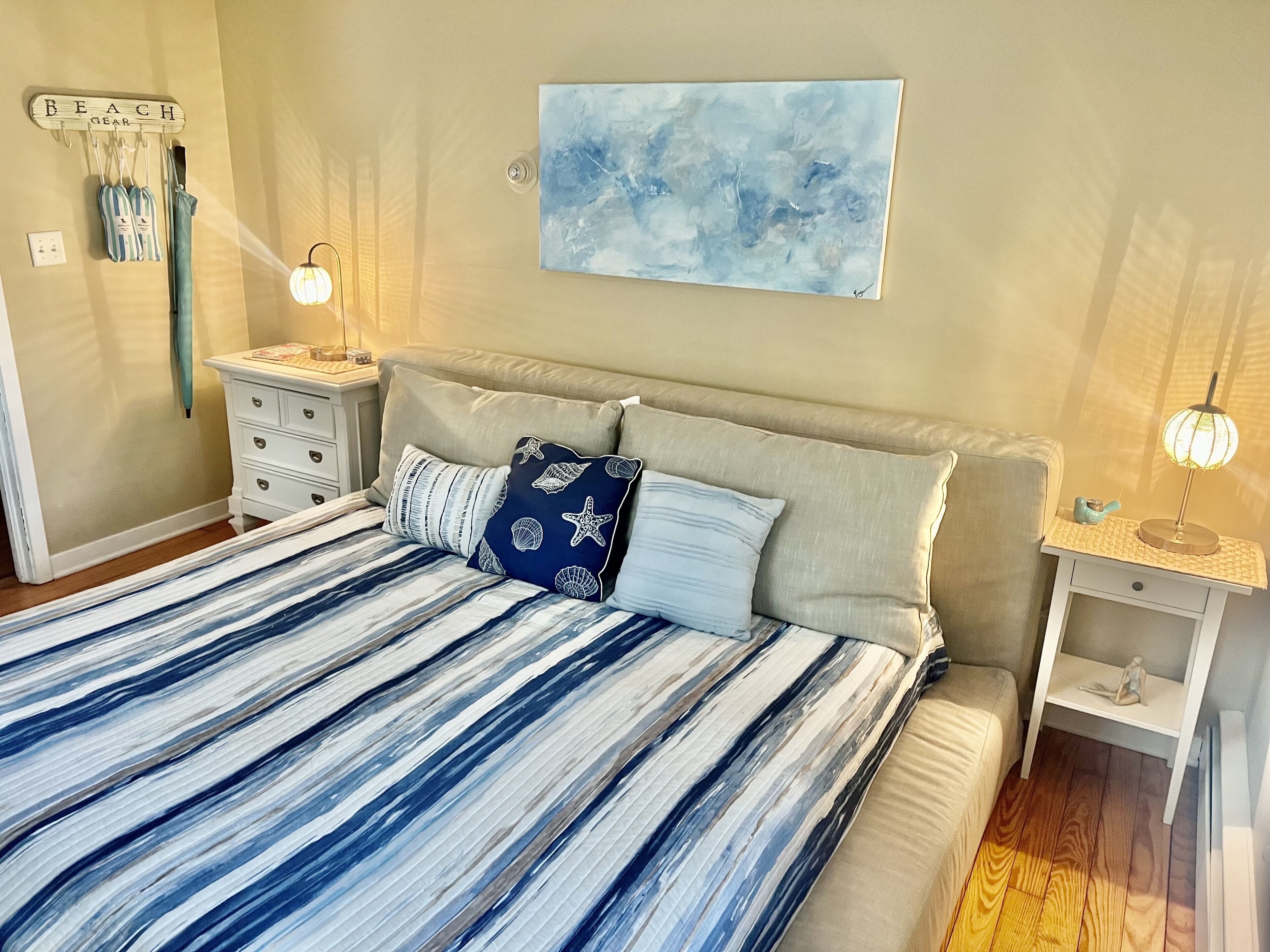 A bedroom with a bed, two nightstands, a wall-mounted shelf, a beach-themed sign, and a painting.