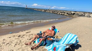 Una spiaggia con una persona sdraiata su una sedia a sdraio, un asciugamano azzurro e bianco a righe, e un borsone blu sulla sabbia.