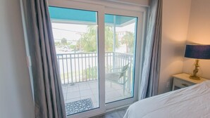 A hotel room with a bed, a nightstand, a lamp, and a view of a balcony with a chair and a railing. - Island Escape 201 Affordable Condo (Clearwater Beach)