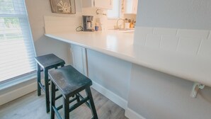 A kitchen with a white countertop, two black stools, a hanging light fixture, and a window with blinds. - Island Escape 201 Affordable Condo (Clearwater Beach)
