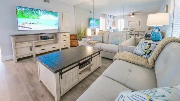 A modern living room with a flat-screen TV, a wooden coffee table, a white sofa, and a dining area in the background. - Island Escape 201 Affordable Condo (Clearwater Beach)