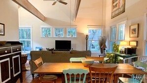 A cozy dining area with a wooden table and chairs, a television in the background, and a window with a view of trees.