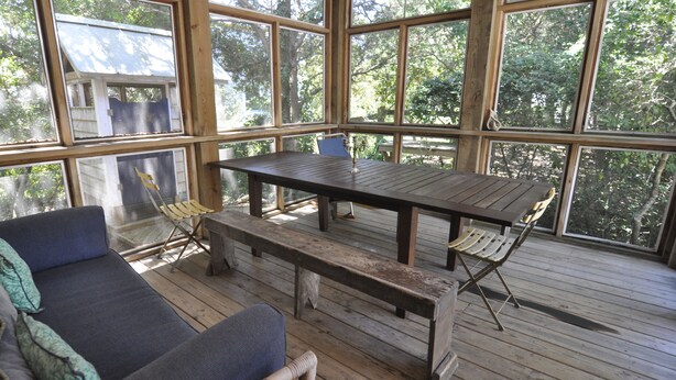 A wooden deck with a table and benches, large windows, and a view of trees.